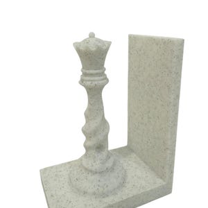 May include: A speckled white chess queen bookend. The queen piece is intricately detailed with a crown and a twisted stem, set on a rectangular base with a vertical support. The bookend is designed to hold books upright.