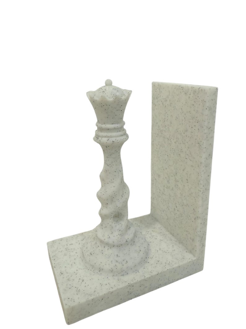 May include: A speckled white chess queen bookend. The queen piece is intricately detailed with a crown and a twisted stem, set on a rectangular base with a vertical support. The bookend is designed to hold books upright.