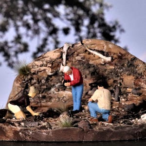 May include: A miniature diorama depicting an archaeological dig site. Three small figures in casual attire are examining the site. The diorama features a rocky outcrop with bone fragments, grass, and a backdrop of trees and a rainbow.