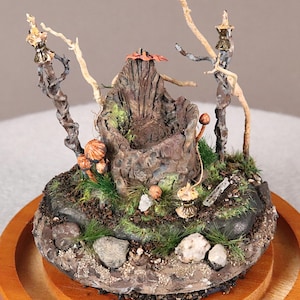 May include: A miniature fairy throne diorama. The throne is carved from a tree stump, surrounded by miniature mushrooms, rocks, and moss. Twig-like structures with gold accents frame the scene, all set on a wooden base.