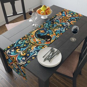 May include: A long rectangular table runner featuring a vibrant, intricate pattern of swirling blue, orange, and yellow floral motifs. A central circular design depicts a black cat silhouette resting inside a golden crescent moon and star.