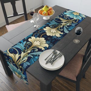 Op de afbeelding: Een lange rechthoekige tafelloper met een herhalend bloemenpatroon met grote crèmekleurige bloemblaadjes, lichtblauwe bladeren en donkerblauwe achtergrondaccenten. De loper ligt op een donkere houten tafel met witte borden en wijnglazen.