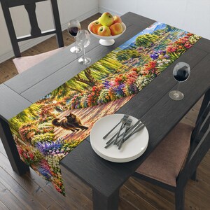 May include: A rectangular table runner featuring a vibrant, colorful garden scene with a black cat walking along a path. The print includes lush green trees, blooming flowers in shades of red, purple, and yellow, and a serene water feature.