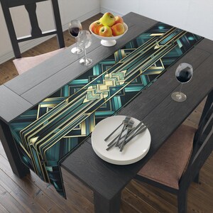 May include: A long rectangular table runner featuring a geometric pattern in shades of teal, black, and gold. The design consists of repeating angular lines and shapes that run the length of the fabric, creating a structured and symmetrical visual effect.