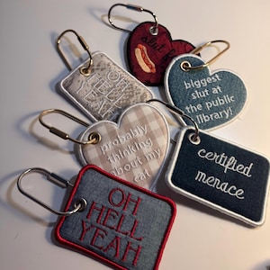 May include: A collection of six embroidered fabric keychains with metal rings. The tags feature various shapes and colors, including text such as OH HELL YEAH, biggest slut at the public library, probably thinking about my cat, and certified menace.