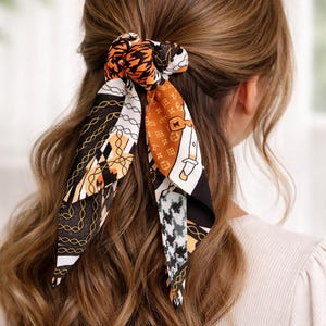 Puede incluir: Una goma para el pelo estampada con un diseño tipo bufanda. La goma presenta una mezcla de patrones negros, blancos y naranjas, incluyendo formas geométricas y eslabones de cadena. El accesorio está atado en el pelo, con los extremos de la bufanda colgando.