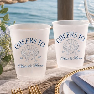 May include: Two frosted plastic cups featuring blue text that reads CHEERS TO, a seashell graphic with two champagne flutes, the year 2027, and the names Olivia & Mason. The cups are positioned on a wooden surface with a blue napkin and gold cutlery.