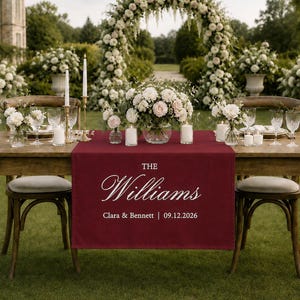 May include: A deep red table runner featuring the white printed text THE Williams Clara & Bennett 09.12.2026, draped over a wooden table with chairs, surrounded by floral arrangements, candles, and a large white flower arch in an outdoor setting.