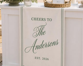 May include: A white fabric table runner featuring a dark green border and the printed text "CHEERS TO The Andersons EST. 2026" in a combination of serif and script fonts, draped over the front of a white rectangular bar counter.