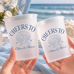 May include: Two frosted plastic cups featuring dark blue text that reads CHEERS TO, with a graphic of a seashell and two clinking champagne flutes. Below the graphic, the text reads 20, 27, and the names Olivia & Mason in a script font.
