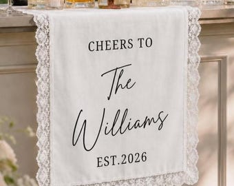 May include: A white fabric table runner with lace trim featuring the black printed text CHEERS TO The Williams EST.2026. The runner is displayed on a bar counter alongside several clear glass decanters filled with amber liquid and crystal glassware.