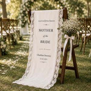 May include: A cream-colored fabric sign draped over a wooden chair, featuring white lace trim. The text reads: reserved from heaven, MOTHER of the BRIDE, Charlene Sweeney, 12.05.2025. A bouquet of baby's breath with a white ribbon is attached.