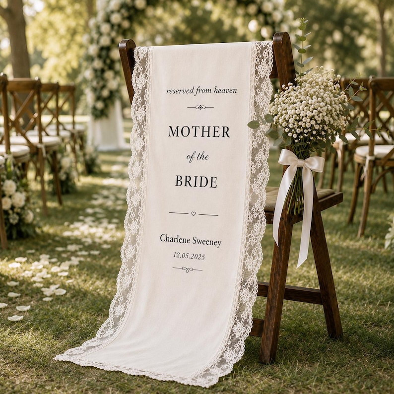 May include: A cream-colored fabric sign draped over a wooden chair, featuring white lace trim. The text reads: reserved from heaven, MOTHER of the BRIDE, Charlene Sweeney, 12.05.2025. A bouquet of baby's breath with a white ribbon is attached.