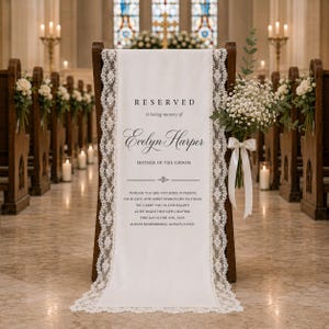May include: A white fabric memorial sign draped over a wooden chair, featuring black text that reads RESERVED in loving memory of Evelyn Harper MOTHER OF THE GROOM, bordered by intricate white lace trim and adorned with a bouquet of baby's breath.