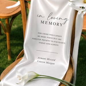 May include: A white satin memorial sign draped over a wooden chair, featuring the black text "in loving MEMORY" followed by "FOR THOSE WHO CANNOT BE HERE TODAY BUT ARE FOREVER PRESENT IN OUR HEARTS, TODAY AND ALWAYS." and the names "Armani Mccord" and "Lauren Morgan".