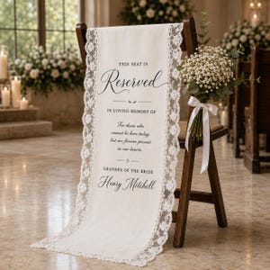 May include: A white fabric memorial sign draped over a wooden chair, featuring lace trim and black text reading: This seat is reserved in loving memory of those who cannot be here today, but are forever present in our hearts. Grandpa of the bride Henry Mitchell.
