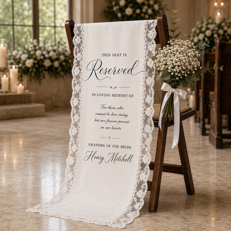 May include: A white fabric memorial sign draped over a wooden chair, featuring lace trim and black text reading: This seat is reserved in loving memory of those who cannot be here today, but are forever present in our hearts. Grandpa of the bride Henry Mitchell.