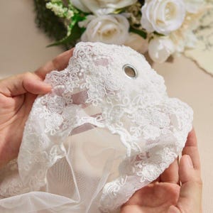 May include: A white lace fabric piece featuring intricate floral embroidery and a scalloped edge. The material includes a single silver-toned metal grommet eyelet near the top center, set against a sheer, lightweight mesh base.