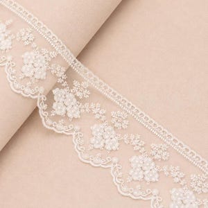 May include: A white lace trim featuring an intricate floral embroidery pattern with a scalloped edge. The delicate design includes small flower clusters and fine thread detailing, creating a decorative border suitable for various textile projects.