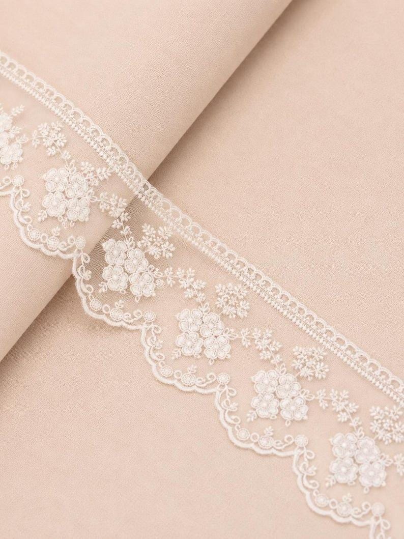 May include: A white lace trim featuring an intricate floral embroidery pattern with a scalloped edge. The delicate design includes small flower clusters and fine thread detailing, creating a decorative border suitable for various textile projects.