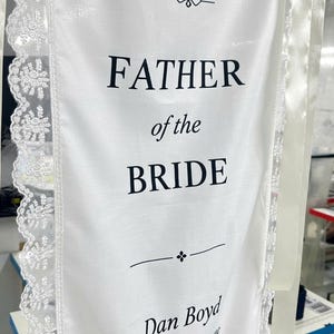 May include: A white fabric banner with lace trim featuring the black text: reserved from heaven, FATHER of the BRIDE, Dan Boyd, and You may not walk me down the aisle, but you'll be with me every step of the way. The banner includes decorative dividers.