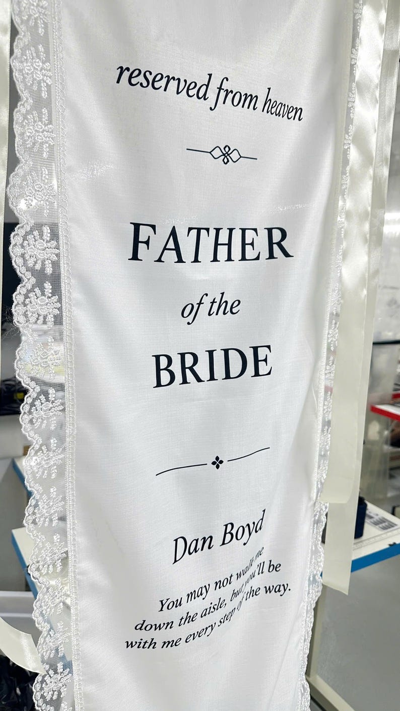 May include: A white fabric banner with lace trim featuring the black text: reserved from heaven, FATHER of the BRIDE, Dan Boyd, and You may not walk me down the aisle, but you'll be with me every step of the way. The banner includes decorative dividers.