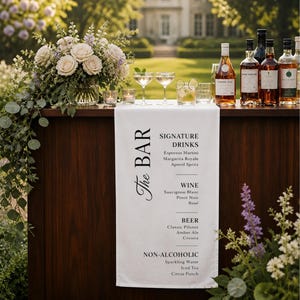 May include: A white fabric bar runner featuring black printed text that reads The Bar, Signature Drinks, Wine, Beer, and Non-Alcoholic, with a list of beverage options under each category, draped over a dark wooden bar counter with bottles.