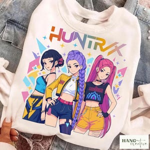 May include: White sweatshirt with a graphic of three anime-style women. The word "HUNTRAX" is printed above the illustration. The women are wearing outfits in blue, yellow, and pink. The sweatshirt has a crew neck. The image is on a denim background.