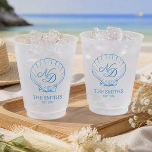 May include: Two frosted plastic cups filled with ice cubes. Each cup features a blue graphic of a seashell with the monogram ND, the text THE SMITHS, and the year EST. 2026 printed in blue ink on the front surface of the clear plastic material.