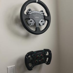 Può includere: Due volanti da corsa neri montati su una parete bianca. Il volante superiore è rotondo con un centro e dettagli argentati. Il volante inferiore è più piccolo, con più pulsanti e quadranti. Il marchio "MOZA" è visibile su entrambi.