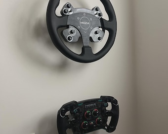 May include: Two black racing steering wheels mounted on a white wall. The top wheel is round with a silver center and accents. The bottom wheel is smaller, with multiple buttons and dials. The brand name "MOZA" is visible on both.