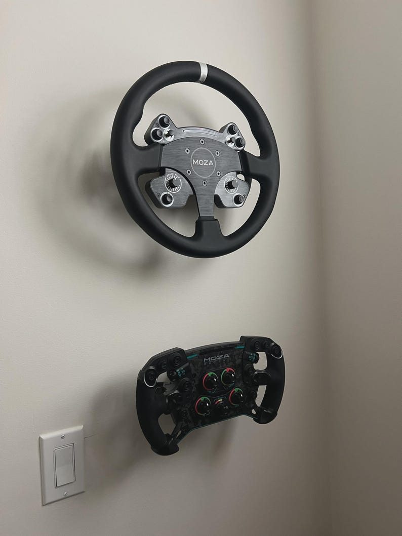 May include: Two black racing steering wheels mounted on a white wall. The top wheel is round with a silver center and accents. The bottom wheel is smaller, with multiple buttons and dials. The brand name "MOZA" is visible on both.