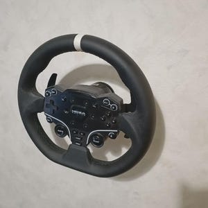 May include: Black racing wheel with a white stripe at the top. The wheel has a black control panel with buttons and the brand name "MOZA". The wheel is designed for use with a racing simulator.