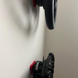 May include: Two black racing steering wheels mounted on a white wall. The wheels are attached to red mounting plates. The word "MOZA" is printed in white on one of the wheels.