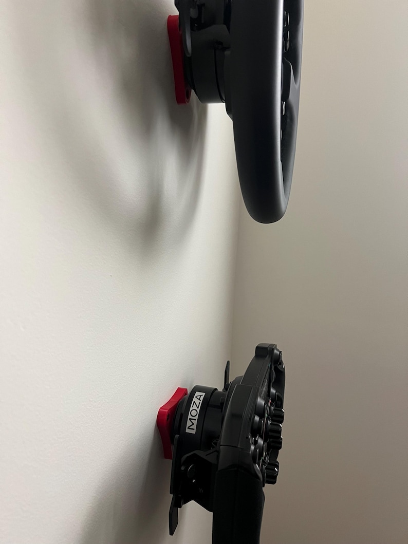May include: Two black racing steering wheels mounted on a white wall. The wheels are attached to red mounting plates. The word "MOZA" is printed in white on one of the wheels.