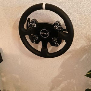 May include: A black steering wheel with a white stripe at the top. The wheel has multiple buttons and dials. The center of the wheel has the word "MOZA" in a white circle. The wheel is mounted on a beige wall.