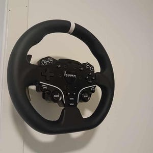 May include: A black steering wheel with a white stripe and a black control panel. The control panel has buttons labeled "RADIO", "HOME", "START", and "MENU". The brand name "MOZA" is visible on the panel. The wheel is mounted on a white wall.