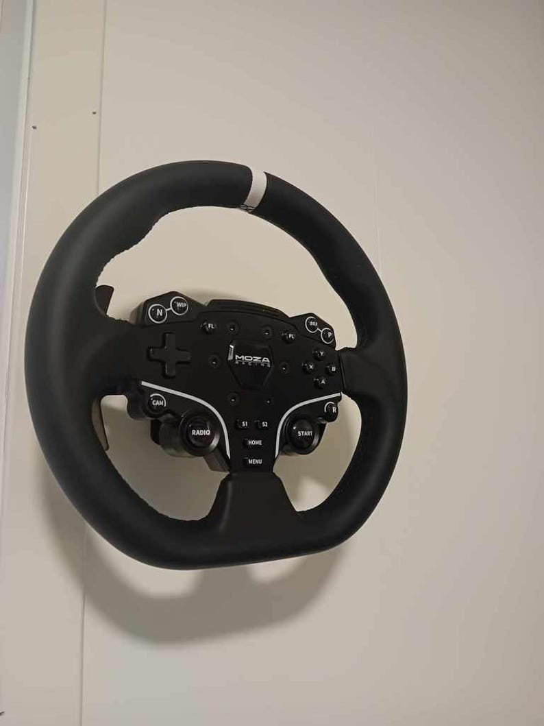 May include: A black steering wheel with a white stripe and a black control panel. The control panel has buttons labeled "RADIO", "HOME", "START", and "MENU". The brand name "MOZA" is visible on the panel. The wheel is mounted on a white wall.