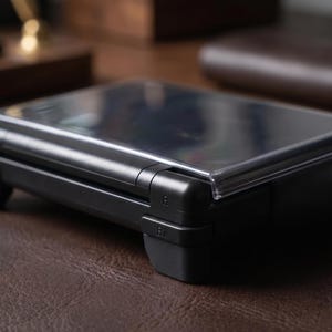 Pode incluir: Um console de jogos portátil preto com um protetor de tela de plástico transparente. O console tem um design elegante com bordas arredondadas e botões visíveis. O dispositivo está sobre uma superfície de couro marrom.