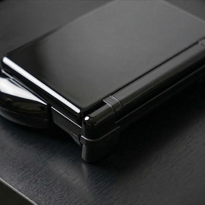 Pode incluir: Um close-up de um console de jogos portátil preto, com um acabamento brilhante. O console está fechado, revelando sua forma retangular e dobradiça. O dispositivo está sobre uma superfície escura, com uma parte da parte inferior do console visível.