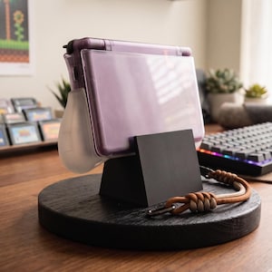 Op de afbeelding: Een paarse handheld gameconsole staat op een zwarte houten standaard met een bruine leren polsband. De standaard staat op een houten bureau, met een toetsenbord en game cartridges op de achtergrond. Een ingelijste afbeelding van een videogame hangt aan de muur.