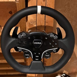May include: Black racing wheel with a white stripe at the top. The wheel has multiple buttons and controls, including "MOZA RACING" text. The wheel is designed for sim racing and features a variety of functions.