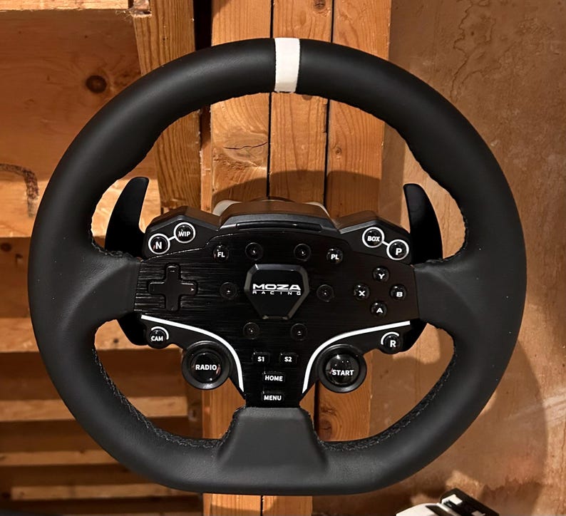 May include: Black racing wheel with a white stripe at the top. The wheel has multiple buttons and controls, including "MOZA RACING" text. The wheel is designed for sim racing and features a variety of functions.