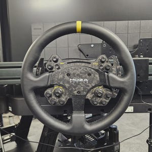 May include: A black racing steering wheel with a yellow stripe at the top. The wheel has multiple buttons and dials, with the "MOZA RACING" logo in the center. The wheel is mounted on a black frame.