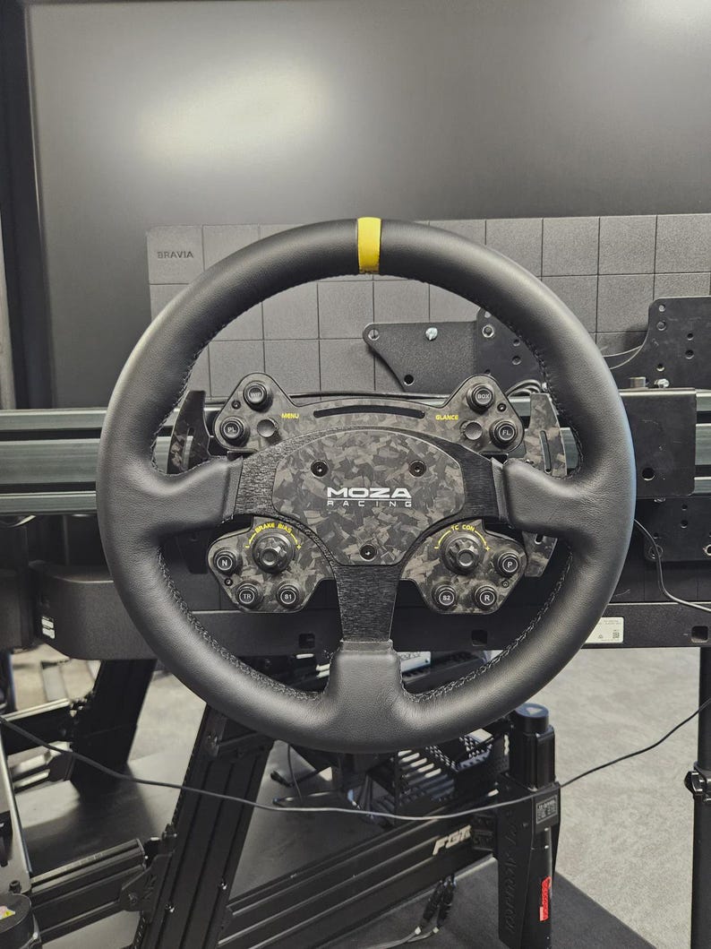 May include: A black racing steering wheel with a yellow stripe at the top. The wheel has multiple buttons and dials, with the "MOZA RACING" logo in the center. The wheel is mounted on a black frame.