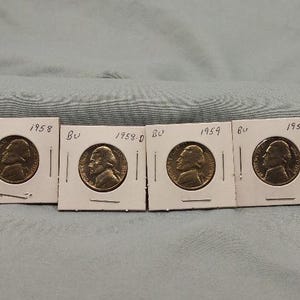 May include: Six vintage US nickel coins, each encased in a small white cardboard holder. The coins are dated 1956, 1958, 1959, and 1945. The coins feature a profile of a man on the obverse.