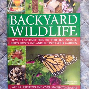 May include: A book titled "Backyard Wildlife" with a green and red cover. The cover features images of a frog, butterfly, bee, and bird, along with other garden wildlife. The book's subtitle reads: "How to attract bees, butterflies, insects, birds, frogs and animals into your garden."
