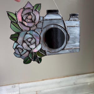 May include: Stained glass camera with a black lens and a floral arrangement of pink and blue roses. The camera is gray and white, with a silver border. The roses have green leaves.