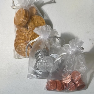 May include: Three sheer white organza drawstring bags filled with round tokens. One bag contains gold-colored tokens, another holds silver-colored tokens, and the third contains copper-colored tokens. Each token features a circular embossed design.