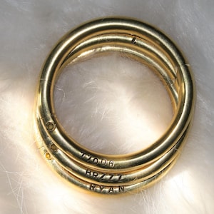 May include: Three gold-colored metal rings stacked together. Each ring has a rounded shape and is engraved with a series of letters and numbers: "77006", "BRZZZ", and "RYAN". The rings are on a white, fluffy surface.